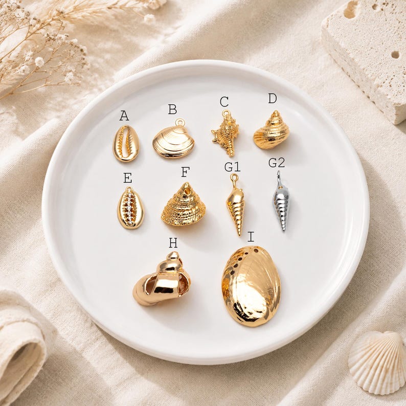 May include: A collection of metal shell-shaped charms labeled A through I, arranged on a white plate. The charms include various gold-toned and one silver-toned seashell designs, each featuring a small loop for attachment, showcasing detailed textures.