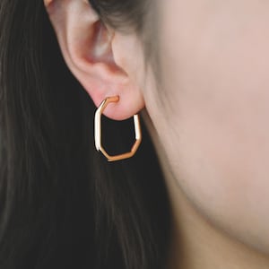May include: Gold octagon hoop earrings. The earrings are worn in a person's ear.