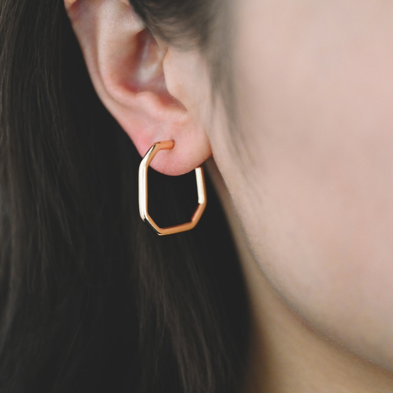 May include: Gold octagon hoop earrings. The earrings are worn in a person's ear.