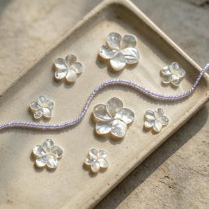May include: A collection of seven white iridescent mother-of-pearl flower-shaped charms arranged on a rectangular beige ceramic tray, accompanied by a single long strand of small, light purple beads that curves across the tray surface.