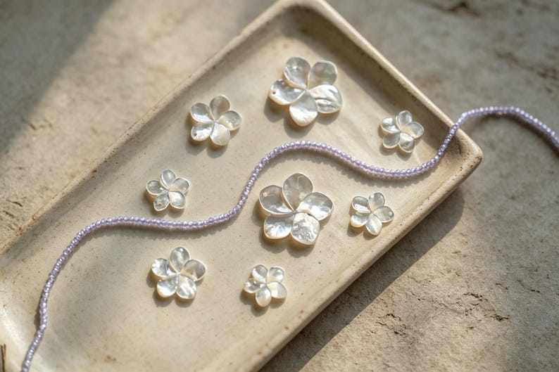 May include: A collection of seven white iridescent mother-of-pearl flower-shaped charms arranged on a rectangular beige ceramic tray, accompanied by a single long strand of small, light purple beads that curves across the tray surface.