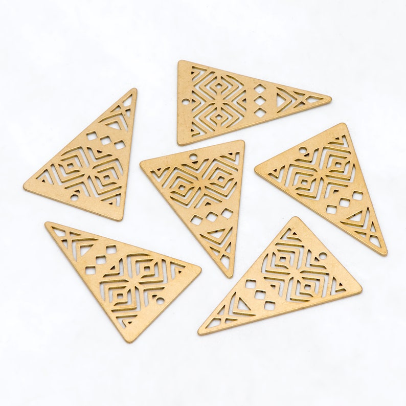 May include: Six gold-colored, triangular metal charms with geometric cut-out designs. Each charm has a small hole at the top for stringing. The charms are arranged on a white surface.
