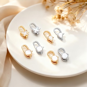 May include: An assortment of jewelry clasps in gold and silver tones, featuring various designs including butterflies, teardrops, and geometric shapes. The clasps have white mother-of-pearl inlays and are arranged on a white plate.