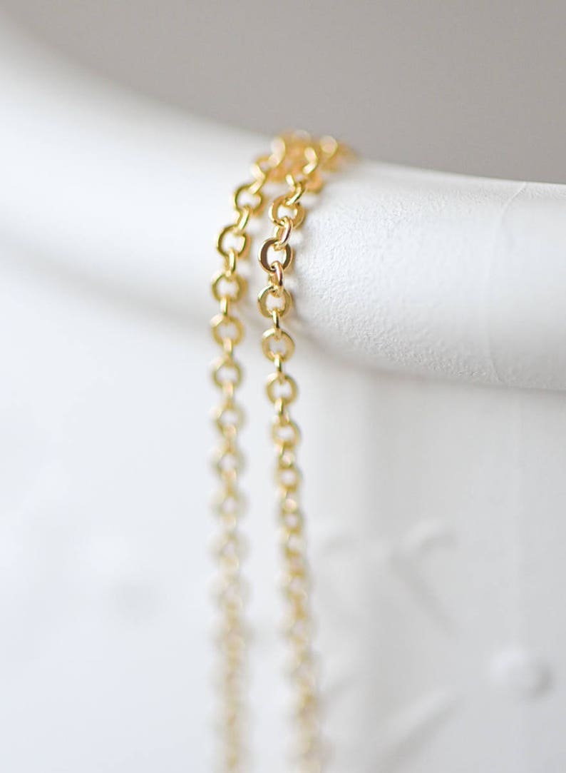 May include: Two gold chains, one slightly thicker than the other, are draped over a white surface. The chains are made of small, linked circles.