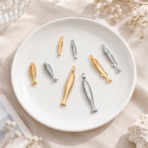 May include: A collection of eight small fish-shaped charms in gold and silver tones, each featuring detailed scale textures and a small loop at the top for attachment. The charms are arranged on a plain white plate, showcasing various sizes.