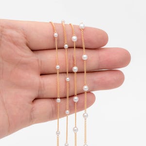 May include: Three gold chains with white pearl beads. The chains are layered on top of each other and are held in a hand.