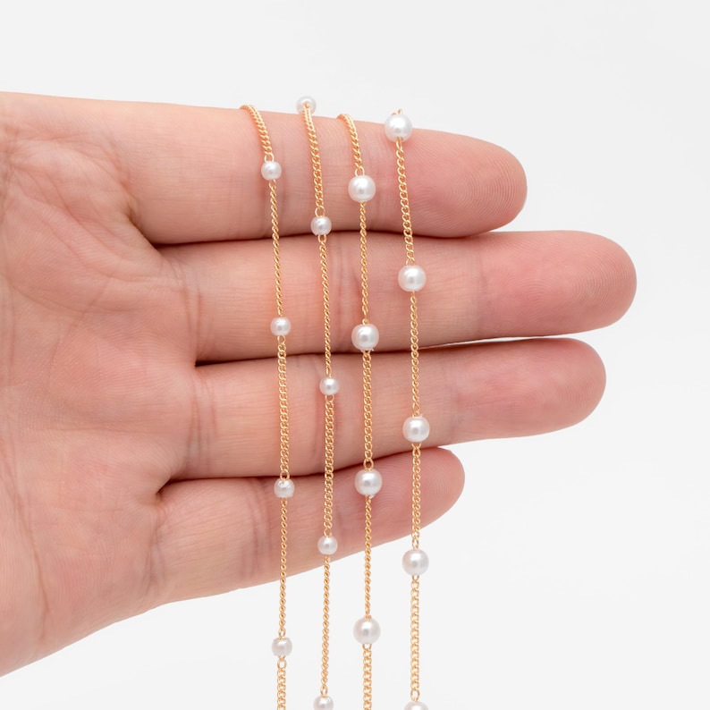 May include: Three gold chains with white pearl beads. The chains are layered on top of each other and are held in a hand.