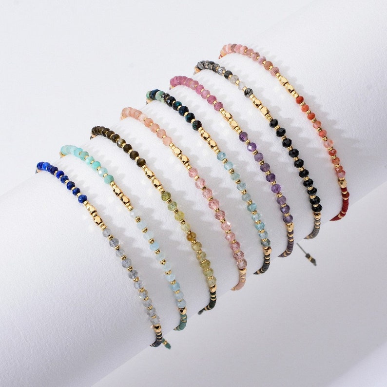 May include: A set of ten beaded bracelets with different colored gemstones, including lapis lazuli, aquamarine, amethyst, and rose quartz. Each bracelet features gold beads and a sliding knot closure.
