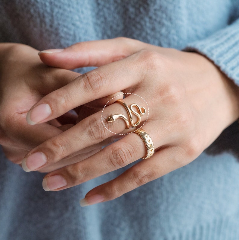 May include: A gold snake ring coiled around a finger, with the snake's head at the top. Another gold ring with a textured design is also visible. The rings are displayed on a hand, with a light blue sweater in the background.