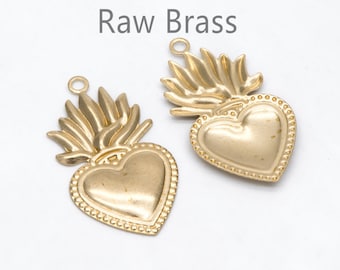 May include: Two raw brass heart-shaped pendants with a flame design above the heart. The pendants have a small loop at the top for attaching to a chain or other jewelry component. The text "Raw Brass" is above the pendants.