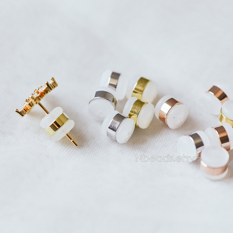 May include: A close-up of a variety of earring backs in gold, silver, and rose gold. The backs are made of metal and have a clear plastic backing. The earring backs are scattered on a white surface.