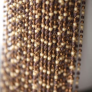 May include: A close-up of a chain made of small, brown and cream colored beads. The chain is made of metal and plastic beads.