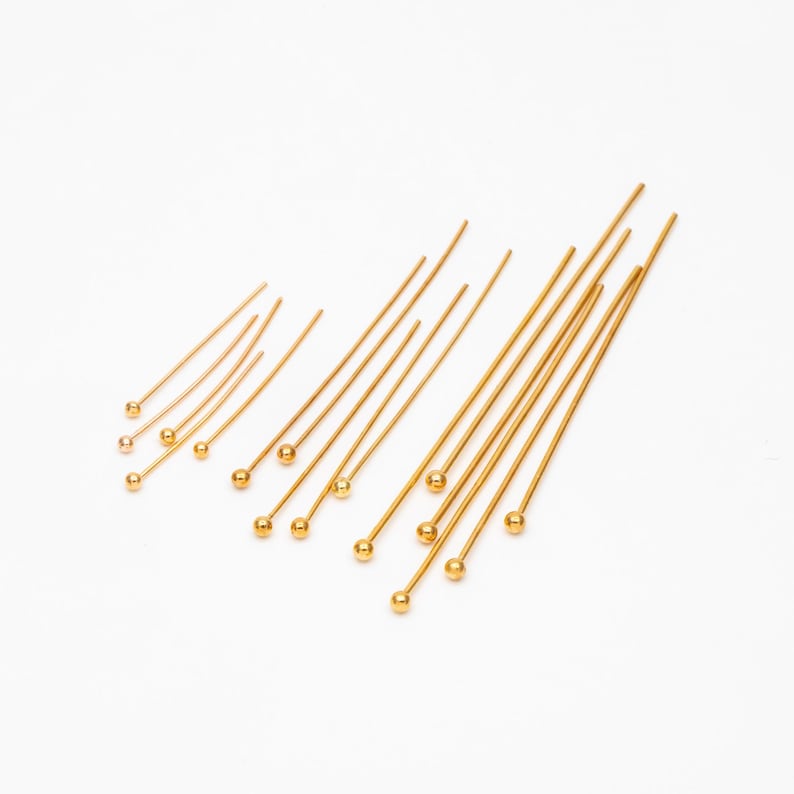May include: A collection of gold head pins of varying lengths, arranged in a fan shape on a white background.