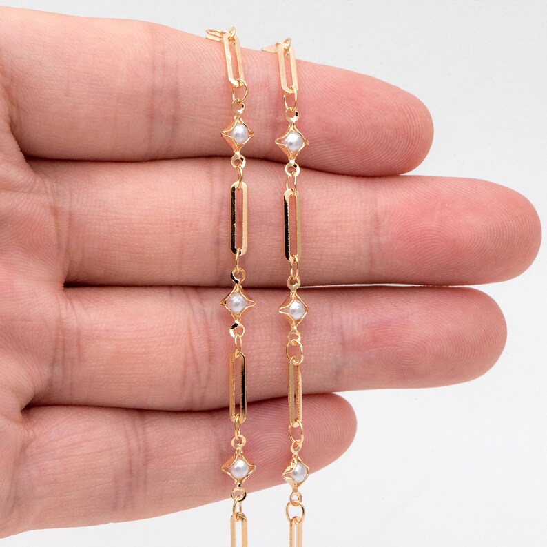 May include: Two gold chains with small white pearls and square-shaped accents. The chains are linked together and are held in a hand.