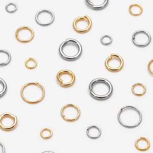 Op de afbeelding: Een verzameling van diverse metalen buigringen in goud- en zilverkleurige tinten, elk voorzien van een kleine opening. Deze ronde sieradenonderdelen variëren in diameter en dikte en zijn willekeurig over het kader verspreid in een patroon.