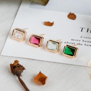 May include: Four pairs of gold stud earrings with square-shaped gemstones. The gemstones are clear, red, green, and light green.