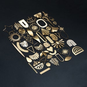 May include: A collection of gold-toned metal charms in various shapes, including stars, suns, hearts, and geometric designs. The charms are arranged in a pattern on a black background.
