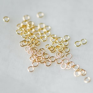 May include: A large pile of gold jump rings, scattered on a white surface. These rings are used for jewelry making and crafting.