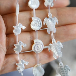 Puede incluir: Hilos de cuentas blancas iridiscentes con formas de vida marina, incluyendo tortugas, conchas marinas y conejos. Las cuentas están ensartadas en un cordón fino y transparente, creando una estética delicada y caprichosa.