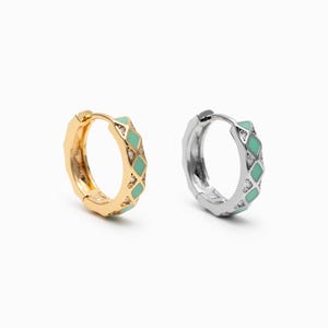 May include: Two hoop earrings are shown, one gold-coloured and one silver-coloured. Both have a geometric pattern with light green enamel and small, clear gemstones. The earrings are set against a white background.