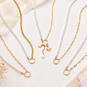 Könnte beinhalten: Eine Sammlung von sechs Halsketten mit gold- und silberfarbenen Ketten, teilweise mit perlenartigen Elementen. Eine zentrale Kette zeigt einen runden Anhänger mit baumelnden Sonnen-, Mond- und Sternmotiven. Jede Kette hat einen runden Verschluss.