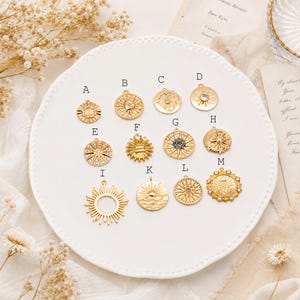 May include: A collection of thirteen gold-toned metal charms labeled A through M, featuring various sun, star, and celestial motifs. Some charms include embedded clear or multicolored crystals, while others display intricate engraved patterns.