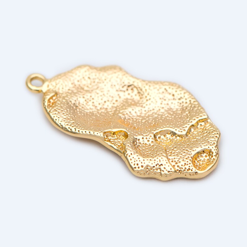 May include: Gold-toned metal pendant with a textured, irregular shape. The pendant has a small loop at the top for attaching to a chain or cord.