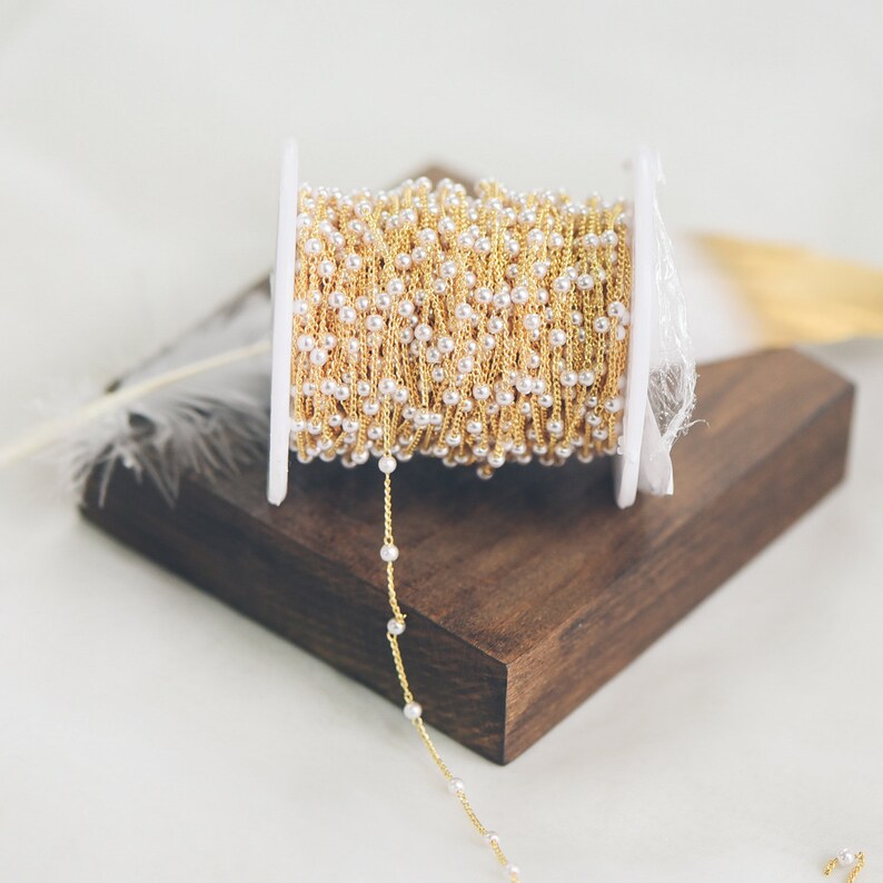 May include: A spool of gold chain with white beads, wrapped around a white plastic spool. The chain is laying on a wooden surface.