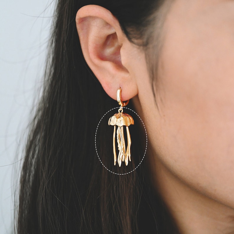 May include: Gold-toned dangle earrings featuring a detailed jellyfish design. The earrings are made of metal and have a hoop style.