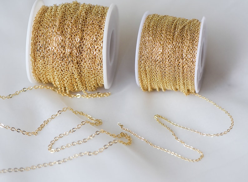 May include: Two spools of gold chain, one larger than the other, with a length of chain extending from each spool. The chain is a delicate, fine link style.