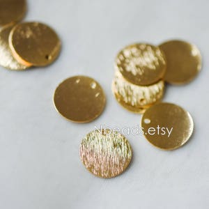 May include: A collection of gold-toned metal round discs with a textured surface and a hole in the center. The discs are arranged in a pile and scattered on a white surface.