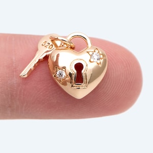 Puede incluir: Un dije de candado en forma de corazón de oro con una cerradura y dos pequeños acentos en forma de estrella con piedras transparentes. Una pequeña llave de oro está unida al candado.
