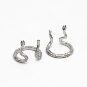 May include: Two silver snake rings with a textured finish. One ring has a snake head with a small clear stone on the top. The other ring has a snake head with a small clear stone on the top.