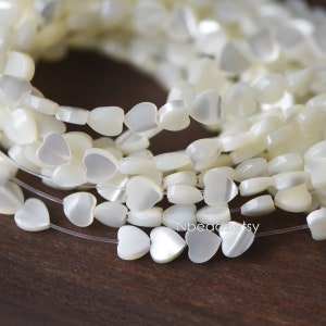 May include: Close-up of strands of white heart-shaped beads. The beads are strung on clear thread, creating a delicate appearance. The beads have a pearlescent finish, reflecting light. The Etsy shop name "Nbeads.etsy" is visible in the image.