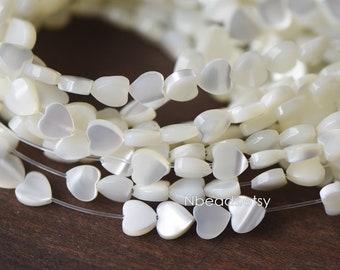 May include: Strands of white heart-shaped beads. The beads are strung together on a clear string, creating a delicate and elegant look. The beads have a pearlescent sheen, reflecting light. The Etsy shop name "Nbeads.etsy" is visible in the image.