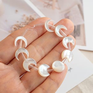 May include: A bracelet featuring white, crescent moon-shaped beads strung together with clear, thin connectors. The beads have a pearlescent sheen, and the bracelet is held in a hand.
