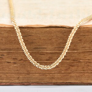 Peut inclure: Un d&eacute;licat collier cha&icirc;ne de couleur or avec un motif textur&eacute; et perl&eacute;. Le collier est pr&eacute;sent&eacute; sur un livre vintage et us&eacute;, de couleur marron chaud. Le motif complexe de la cha&icirc;ne est clairement visible sur la surface vieillie du livre.