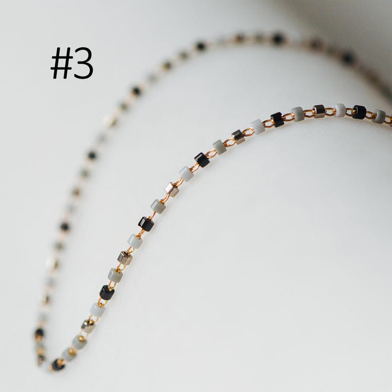 May include: A delicate gold chain necklace with small square-shaped beads in black, gray, and white.