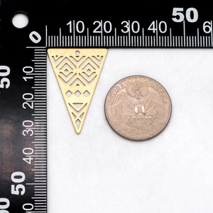 May include: A gold-colored, laser-cut metal triangle pendant with geometric patterns. The pendant is next to a US quarter for size comparison. The pendant has a small hole at the top for stringing.
