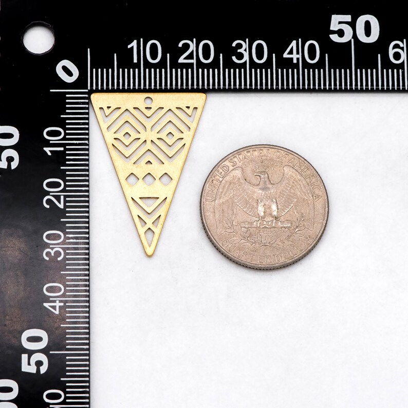 May include: A gold-colored, laser-cut metal triangle pendant with geometric patterns. The pendant is next to a US quarter for size comparison. The pendant has a small hole at the top for stringing.
