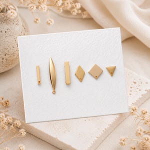 May include: A set of six gold-toned metal jewelry charms arranged on a white display card. The collection includes a thin rectangular bar, a long tapered leaf shape, a wider rectangular bar, a diamond shape, a square, and a small triangle.