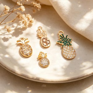 May include: Four gold-colored pineapple charms with varying designs and sizes. Some are encrusted with small, clear crystals, while others have openwork patterns. One charm features green accents on the pineapple crown. The charms are displayed on a white plate.