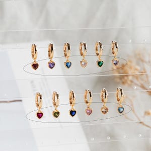Puede incluir: Un conjunto de 10 pendientes de aro dorados con un colgante de corazón. Cada colgante de corazón tiene una gema de color diferente: rojo, morado, azul, verde, rosa, azul claro, azul oscuro y transparente.