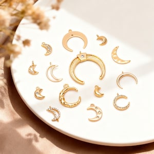 May include: A collection of gold-colored crescent moon charms in various sizes and designs. Some moons are smooth, others textured, and some are embellished with crystals or stars. The charms are arranged on a white plate.