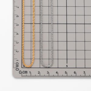 Peut inclure: Deux colliers d&eacute;licats, l'un dor&eacute; et l'autre argent&eacute;, sont pr&eacute;sent&eacute;s c&ocirc;te &agrave; c&ocirc;te sur un fond quadrill&eacute; avec une r&egrave;gle. Les cha&icirc;nes ont un design &agrave; maillons carr&eacute;s. La r&egrave;gle affiche des mesures en centim&egrave;tres.