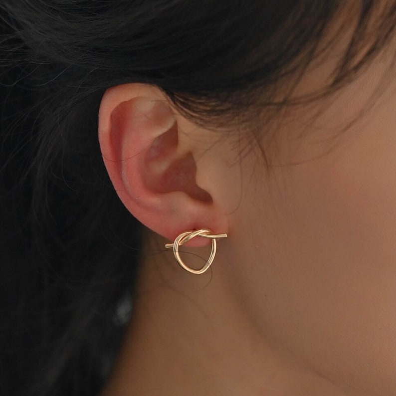 May include: Gold knot-shaped earrings with a small bar extending from the knot.
