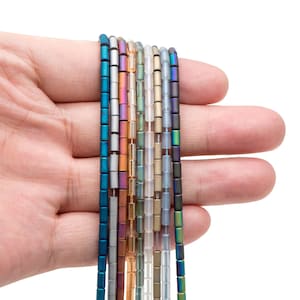 Puede incluir: Una colección de 10 hilos de cuentas cuadradas en varios colores iridiscentes, incluyendo azul, verde, morado, plateado y dorado. Las cuentas están enhebradas en un hilo transparente y se sostienen en una mano.