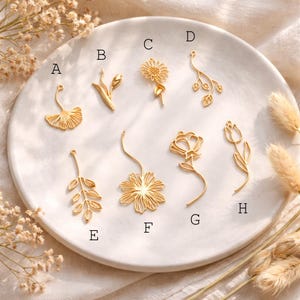 May include: A collection of eight gold-colored floral charms, including a ginkgo leaf, tulip, sunflower, and various other flower and leaf designs. The charms are arranged on a white, circular plate, showcasing their intricate details and delicate forms.