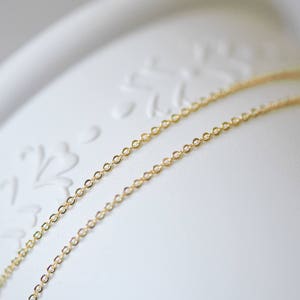 May include: Two gold chains, one on top of the other, lay on a white surface with a floral design.