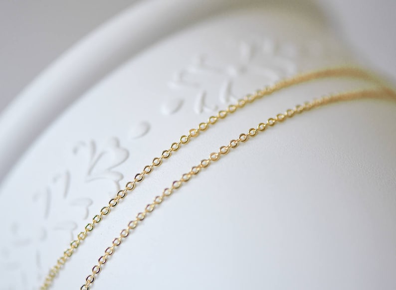 May include: Two gold chains, one on top of the other, lay on a white surface with a floral design.